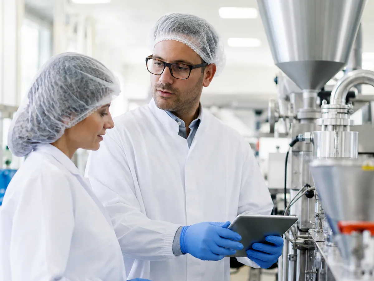 An auditor in a white coat and hairnet reviewing a tablet with a plant worker beside stainless production equipment